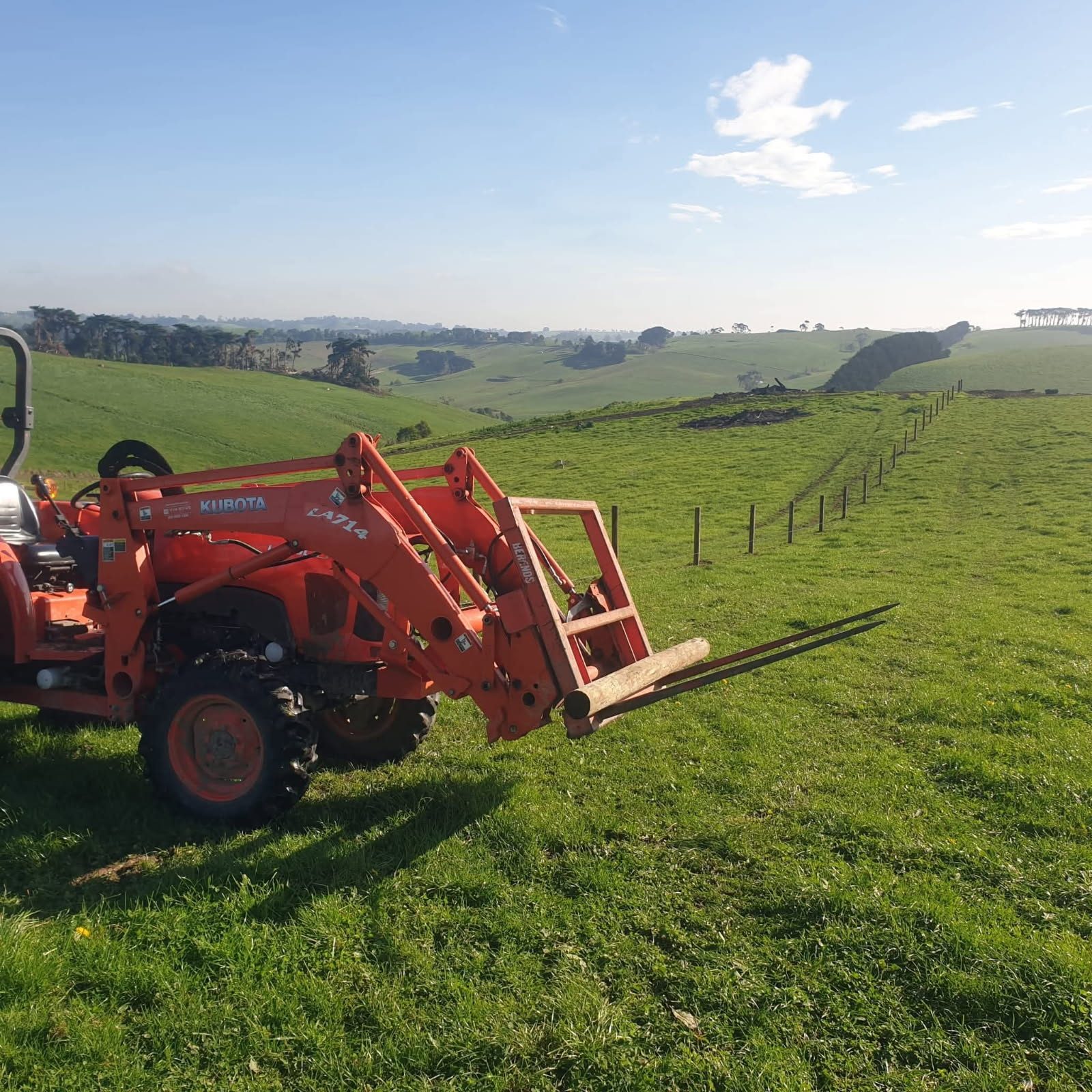 Tractor with pallet forks carrying a fence post across a hillside paddock