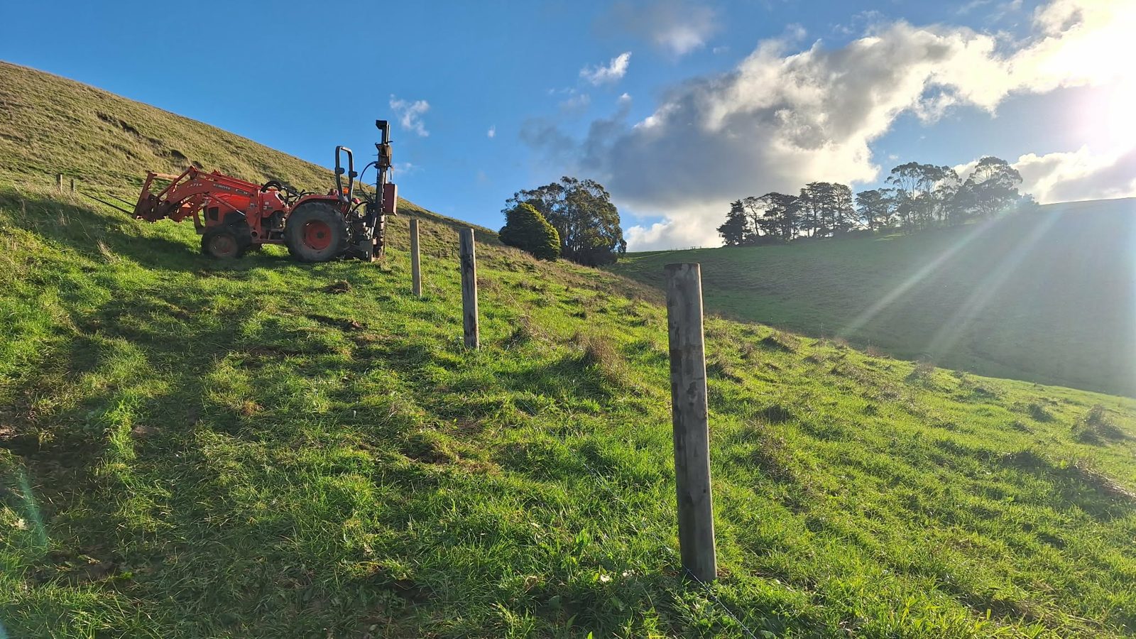 Tractor and post driver on a sunlit hillside paddock with new fence posts
